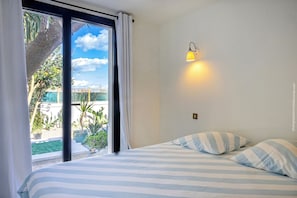 A bedroom with a bed, striped pillows, a nightstand, a wall-mounted lamp, and a view of a pool and greenery outside.