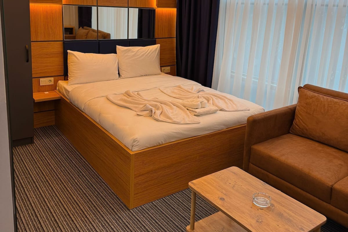 A modern hotel room with a large bed, a wooden headboard, a wooden nightstand, a wooden coffee table, and a brown sofa.