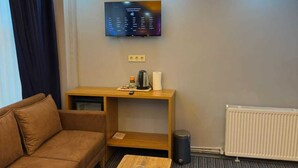 Een moderne hotelkamer met een bank, een houten salontafel, een flatscreen televisie aan de muur, een klein tafeltje met waterkoker en beker, en een radiator op de vloer.