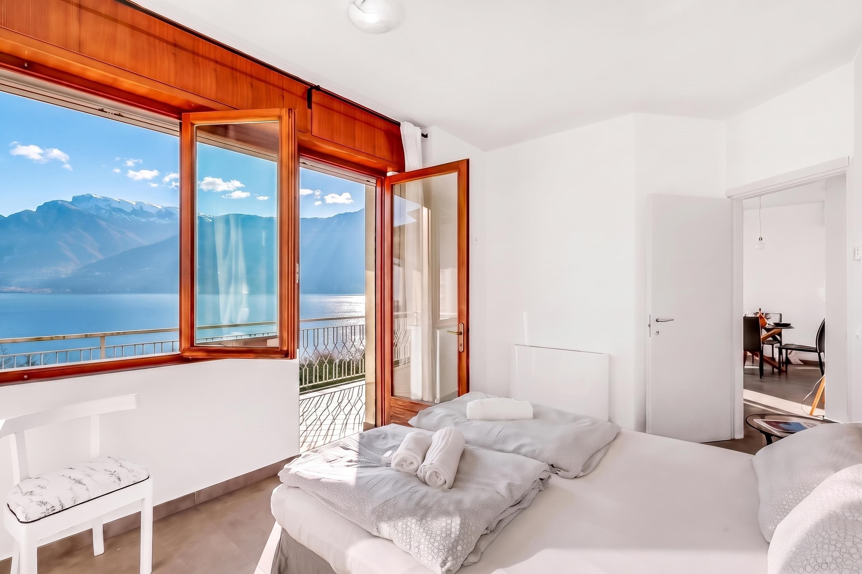 Ein Schlafzimmer mit großem Fenster, das Blick auf Berge und See bietet, ein Bett mit Kissen, ein Sessel und ein kleiner Tisch mit Lampe.