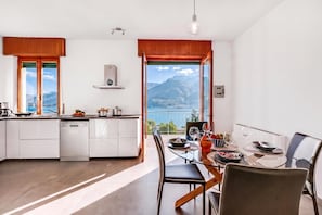 A modern kitchen with a view of mountains, a dining area with a table set for two, and a large window. - Casa Panorama in Tremosine With View Terrace (Tremosine)