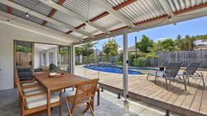 Eine überdachte Terrasse mit Holztisch und -stühlen, einem Pool und Blick auf den Garten.