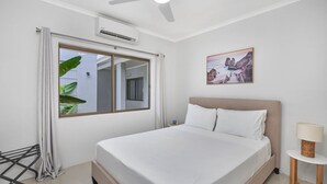 Ein Hotelzimmer mit Bett, einem Vorhangfenster, einem Deckenventilator, einer Klimaanlage, einem gerahmten Bild an der Wand und einem Nachttisch mit Lampe.