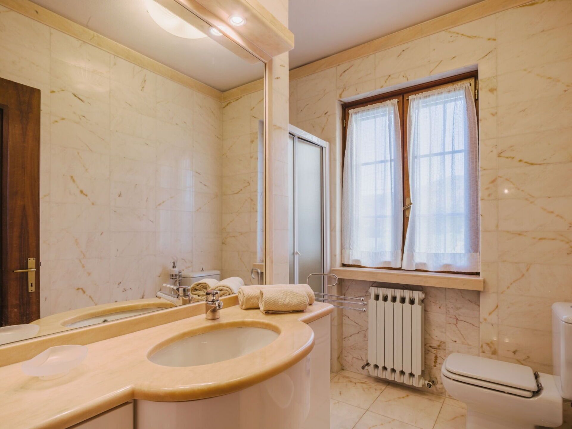 Ein Badezimmer mit Waschbecken, Toilette, Fenster mit Vorhängen und Heizkörper.