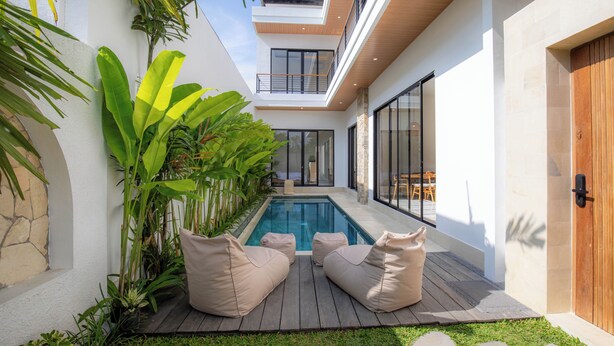 A modern outdoor pool area with a wooden deck, two beige cushioned chairs, and lush tropical plants. - Villa Mallorca Ubud (Ubud)