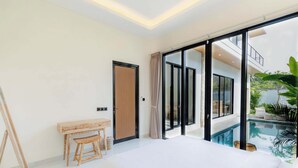 A modern bedroom with a large bed, a wooden desk, and a view of a swimming pool and outdoor area. - Villa Mallorca Ubud (Ubud)