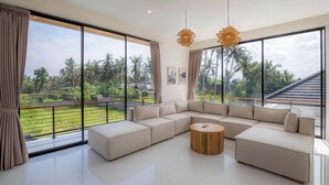 A modern living room with a large sectional sofa, a round wooden coffee table, and large windows offering a view of a lush garden and palm trees. - Villa Mallorca Ubud (Ubud)