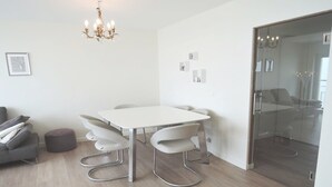 Una habitación moderna con una mesa y sillas blancas, un sofá gris, un pequeño reposapiés y un separador de vidrio.