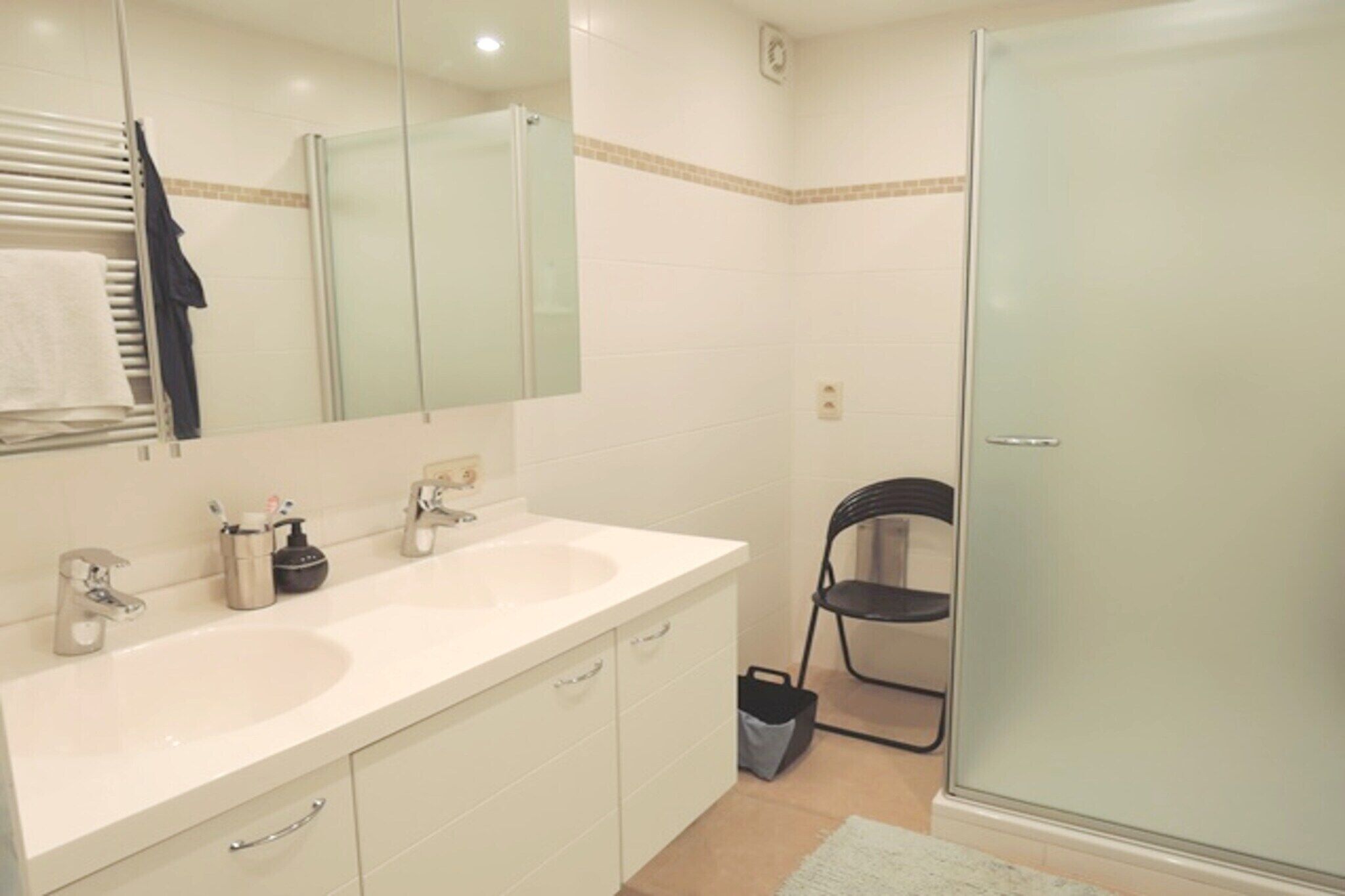 A bathroom with two sinks, a mirror, a towel rack, a chair, and a glass shower enclosure.