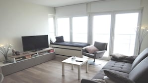 Una sala moderna con un sofá, una mesa de centro, un televisor en un mueble bajo y un asiento junto a la ventana con vistas.
