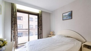 A hotel room with a large bed, a bedside table, a lamp, a window with curtains, and a painting on the wall.