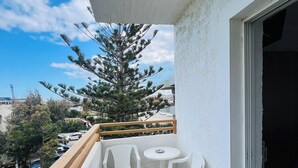 A balcony with a white railing, a small table with two chairs, and a view of a pine tree and distant buildings. - Mark Hotel (Heraklion)