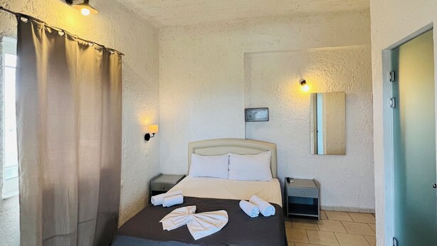 A bedroom with a bed, a nightstand, a wall-mounted lamp, a door, and a window with curtains. - Mark Hotel (Heraklion)