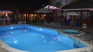 An outdoor swimming pool with a tiled edge, surrounded by lounge chairs and umbrellas, and a bar area with people. - Mark Hotel (Heraklion)
