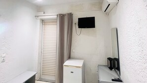 A small, compact room with a white air conditioner, a small refrigerator, a bench, a desk with a mirror, and a window with curtains. - Mark Hotel (Heraklion)