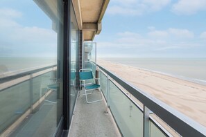 A balcony with a glass railing overlooking a beach. There are two green chairs and a small table.