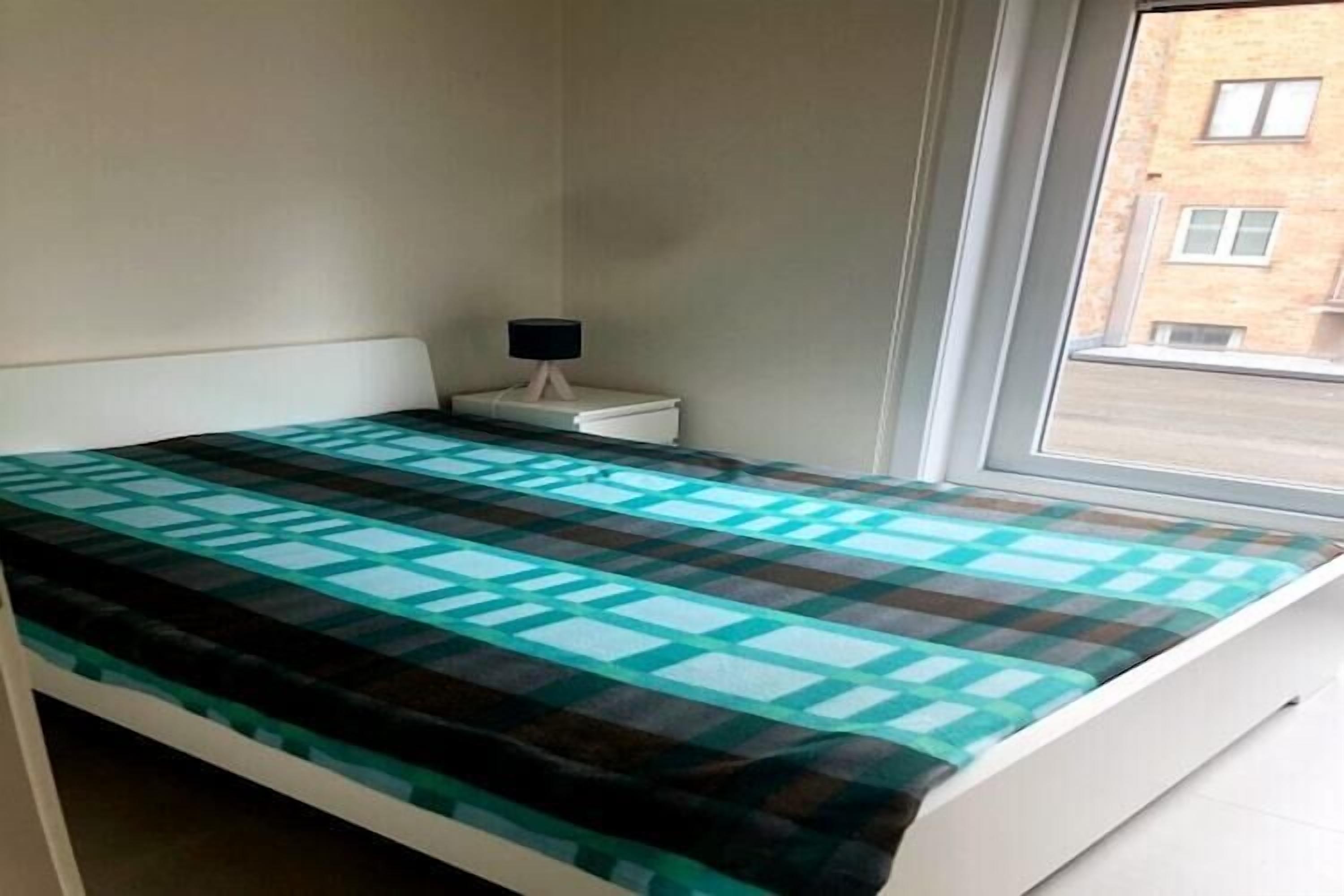 A single bed with a checkered bedspread, a bedside table with a lamp, and a window with a view of buildings.