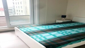 Ein Schlafzimmer mit Bett, Nachttisch, Lampe und Fenster mit Blick auf Gebäude.