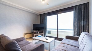 A modern hotel room with two sofas, a small table, a TV, and a large window with curtains. - Apartment in Middelkerke near Beach & Casino (MIDDELKERKE)