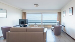 Una sala de estar moderna con un sofá grande, un televisor de pantalla plana y vistas al océano a través de ventanas amplias.