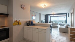 A modern kitchen with white cabinets, a built-in oven, and a coffee maker. There is a dining area with a white table and chairs, and a large window offering a view of the ocean. - Apartment in Middelkerke near the Beach (MIDDELKERKE)