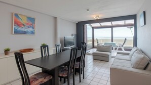 A modern living room with a dining area, a flat-screen TV, and a large window overlooking the sea. - Apartment in Middelkerke near the Beach (MIDDELKERKE)