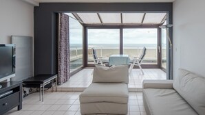 A modern living room with a large window overlooking the sea, a flat-screen TV, a sofa, and a small table. - Apartment in Middelkerke near the Beach (MIDDELKERKE)