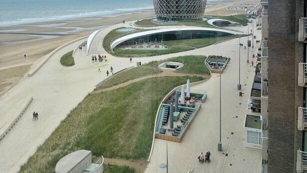 Un bâtiment moderne au design sphérique unique, situé près d’une plage, avec un cheminement piétonnier incurvé et des espaces aménagés pour s’asseoir.