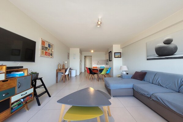 A modern living room with a flat-screen TV, a sofa, a dining table with colorful chairs, and a kitchen in the background. - Apartment in Nieuwpoort with Stunning Sea Views (MIDDELKERKE)