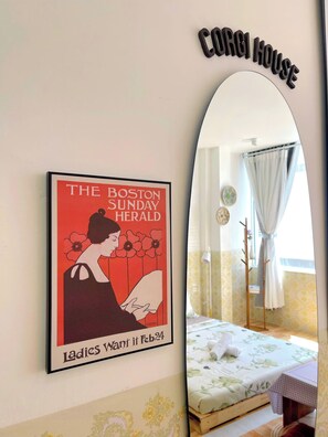A framed poster of 'The Boston Sunday Herald' with a date 'Feb 24' and a room with a bed, a table, and a mirror reflecting a window with curtains.
