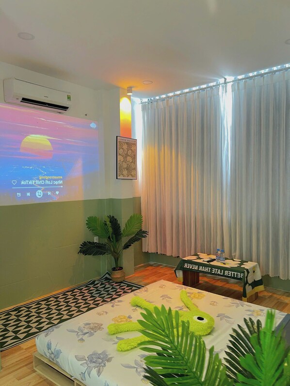 A hotel room with a bed, a projector screen displaying a movie, a potted plant, a table with a water bottle, and a curtain.