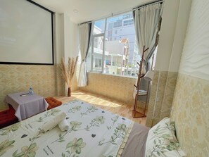 A hotel room with a bed, a small table with a water bottle, a vase with pampas grass, and a window with curtains.