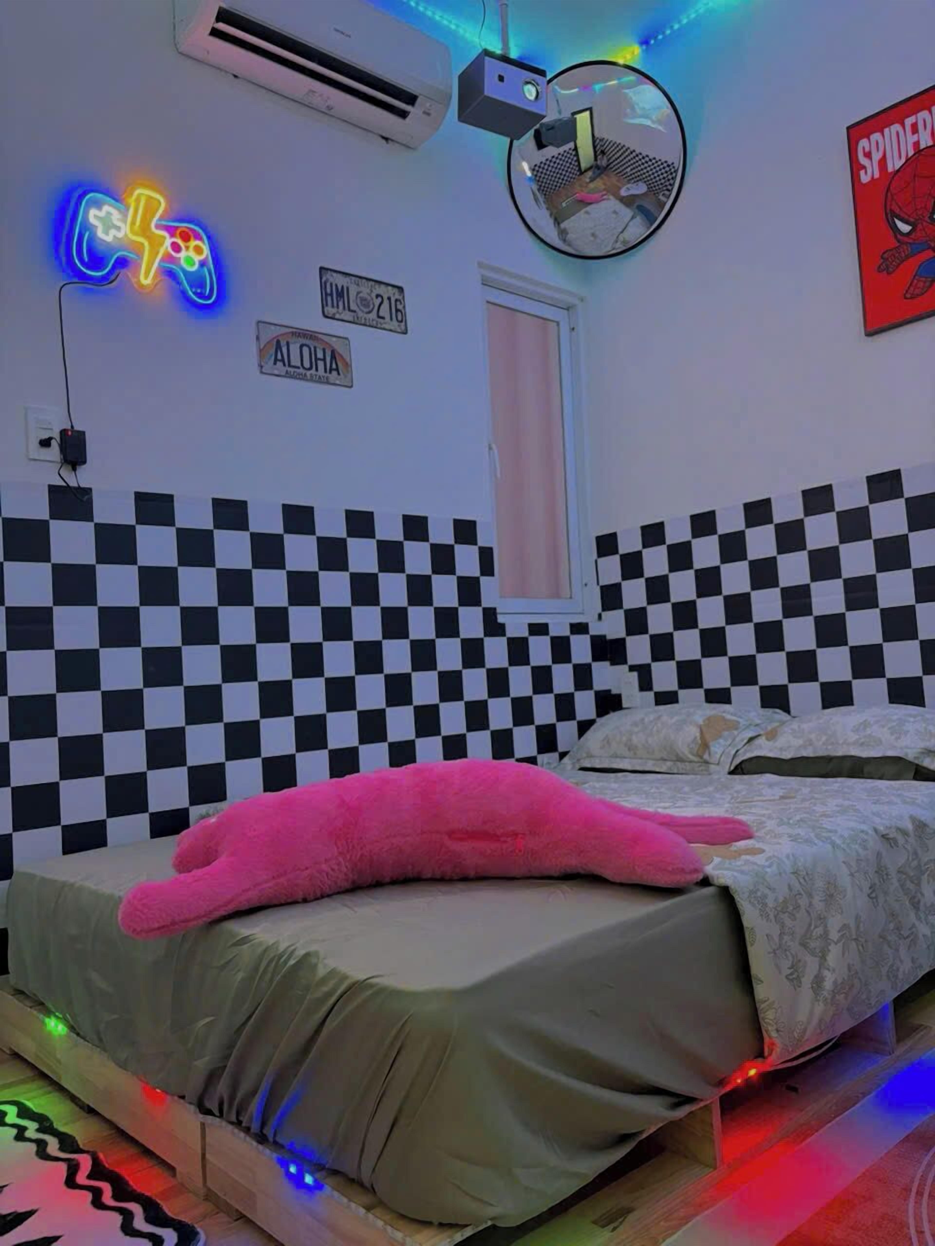 A bedroom with a checkered wall, a neon sign, a Spiderman poster, and a bed with a pink pillow.