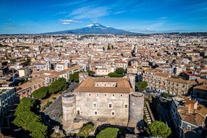 A historic cityscape with a prominent castle-like structure surrounded by dense urban buildings, a clear sky, and a distant mountain. - Fiera House in the City Center (Catania)