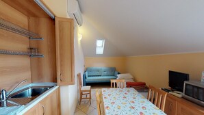 Eine kompakte Küchenzeile mit Spüle, Schränken und einer Mikrowelle. Ein kleiner Essbereich mit Tisch und Stühlen lädt zum Verweilen ein, im Hintergrund befindet sich eine Couch.