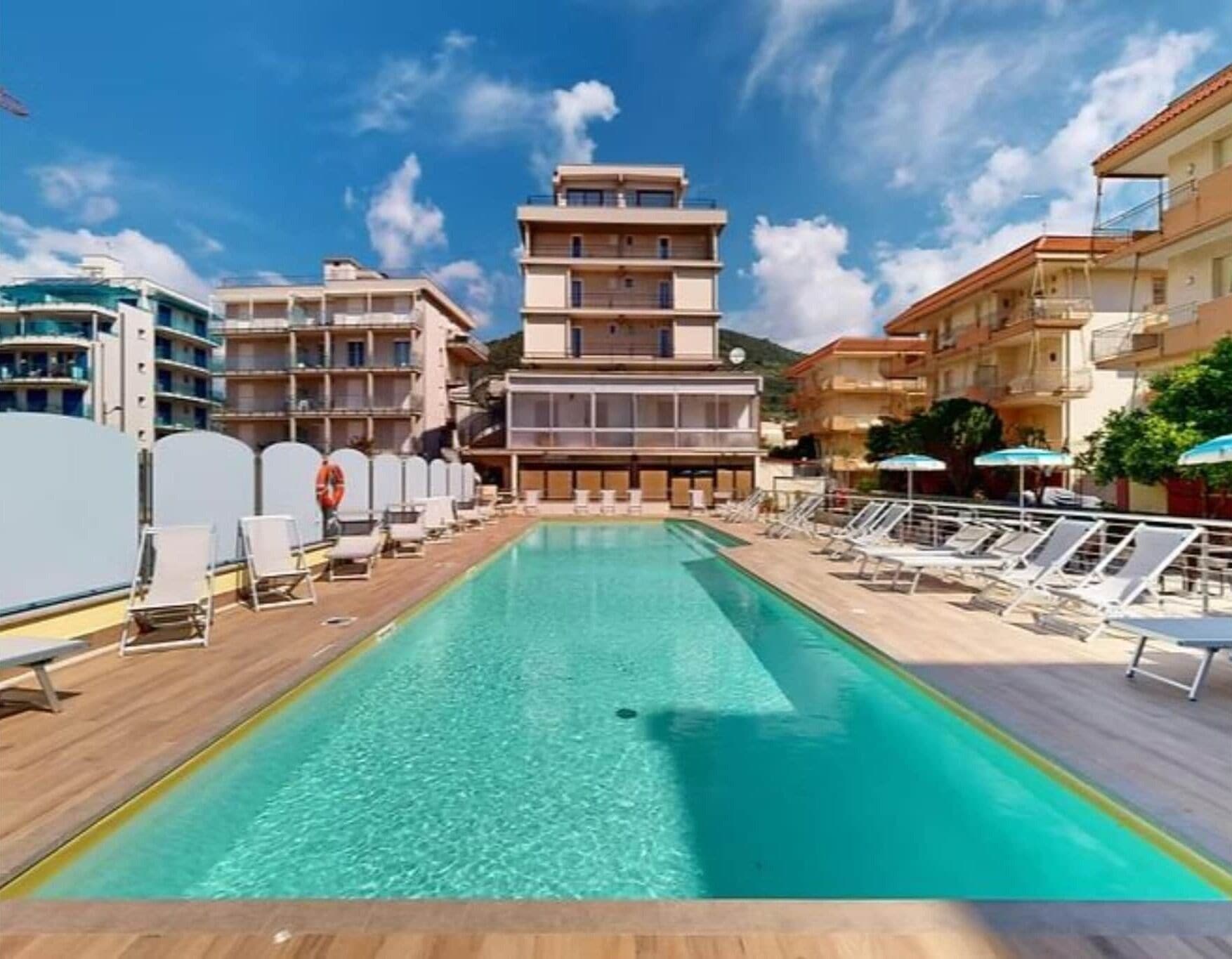 Zona de piscina con agua cristalina, rodeada de tumbonas y sombrillas, con edificios al fondo.