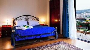 A neatly made bed with a blue bedspread and white pillows, a wooden floor, a nightstand with a lamp, and a view of a balcony with outdoor seating. - AIGES MELATHRON (Veroia)