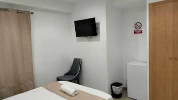 Habitación de hotel con cama, televisión montada en la pared, una silla, un refrigerador pequeño y un cesto de basura.