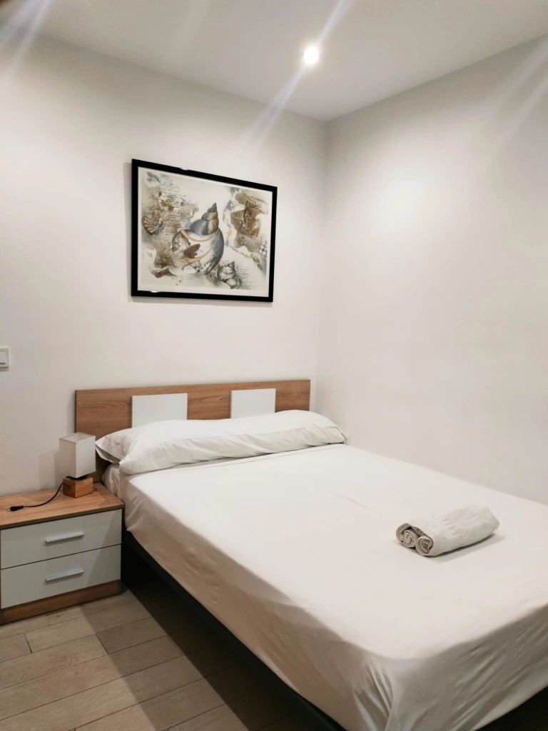 Een keurig opgemaakt slaapkamer met een bed, nachtkastje met lamp, lade en een lijst met kunstwerk aan de muur.