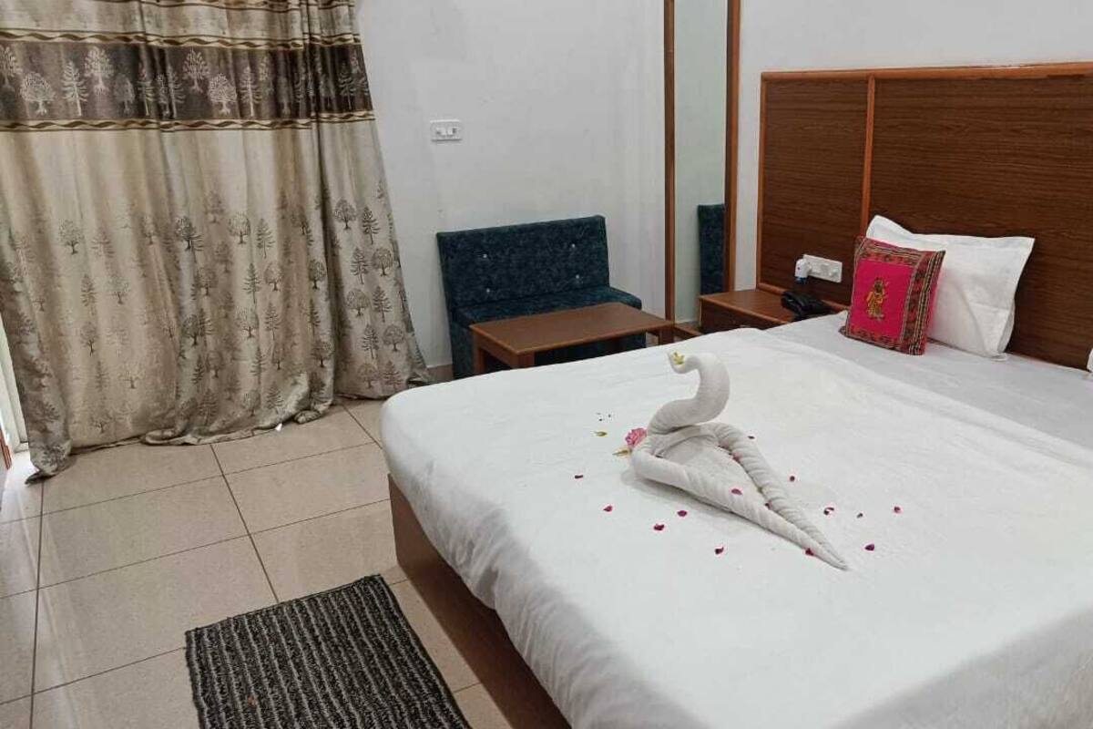 A hotel room with a large bed, a wooden headboard, a small table, a blue chair, and a curtain with a tree pattern.