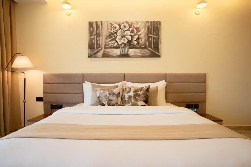 A hotel room with a large bed, a headboard, two bedside lamps, a nightstand, and a wall-mounted painting.