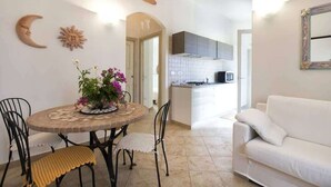 A dining area with a round table, chairs, and a flower centerpiece. There is a white sofa and a kitchen in the background. - Casa 10 Da Georgette - Affitti Brevi Italia (Valledoria)