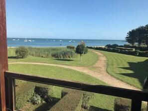 Une vue depuis un balcon donnant sur un espace verdoyant, avec un sentier menant à une plage où l’on aperçoit des bateaux au loin.