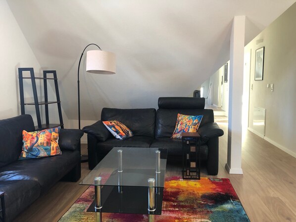 Un salon avec un canapé en cuir noir, des coussins colorés, une table basse en verre, un lampadaire et un tapis aux couleurs vives.