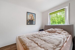 Un dormitorio con cama, ventana con vista a árboles y un cuadro en la pared.