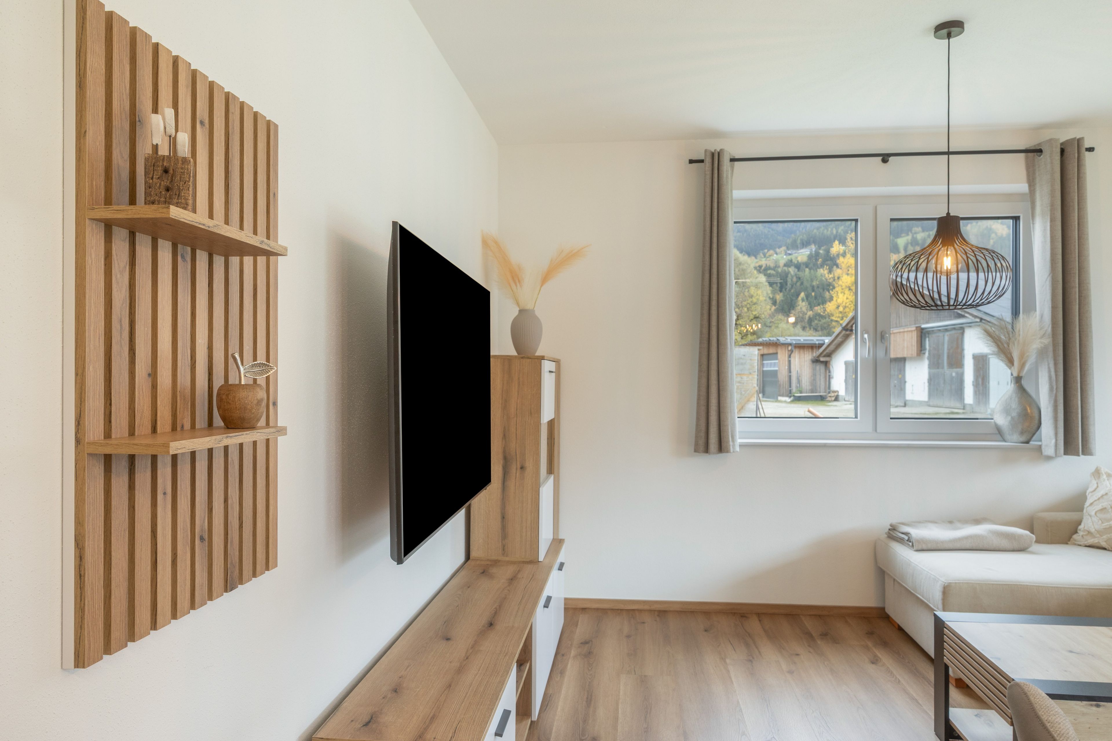 Ein modernes Wohnzimmer mit Flachbildfernseher an einer Holzvertäfelung, einer Couch, einem Holztisch und einem Fenster mit Blick auf ein Haus und Bäume.
