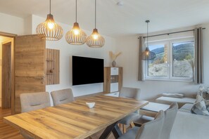 Una sala de estar moderna con una mesa de comedor de madera, sillas grises tapizadas, un televisor de pantalla plana montado en la pared y una ventana con vistas a un paisaje montañoso.