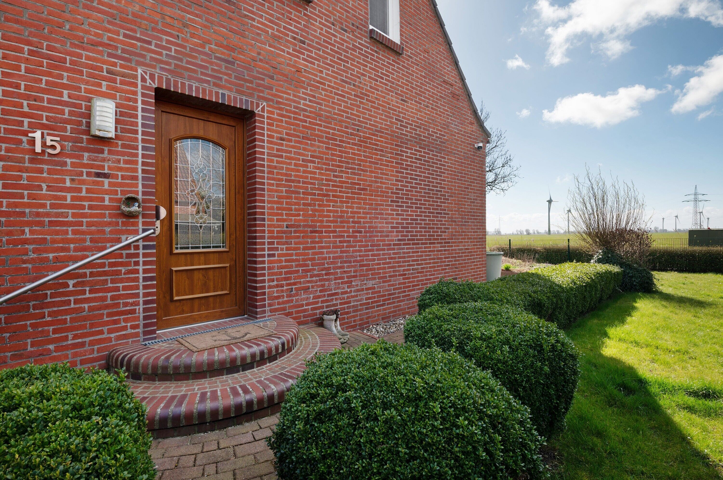 A red brick building with a wooden door, numbered 15, and a small garden with hedges.