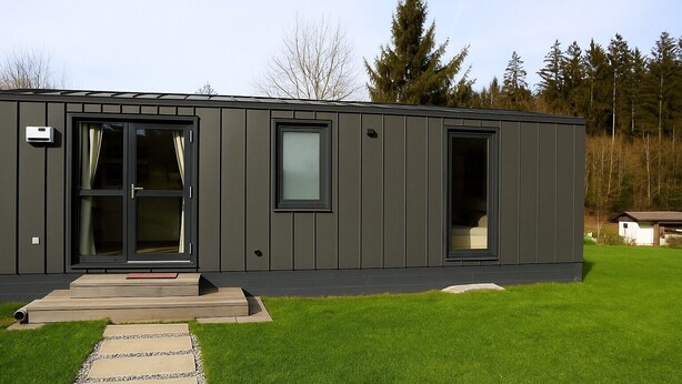 A modern, single-story building with a dark exterior, large windows, and a well-maintained lawn. - Tiny Home 'Seppl' with Mountain View, Wi-Fi, and Air Conditioning (Untergriesbach)