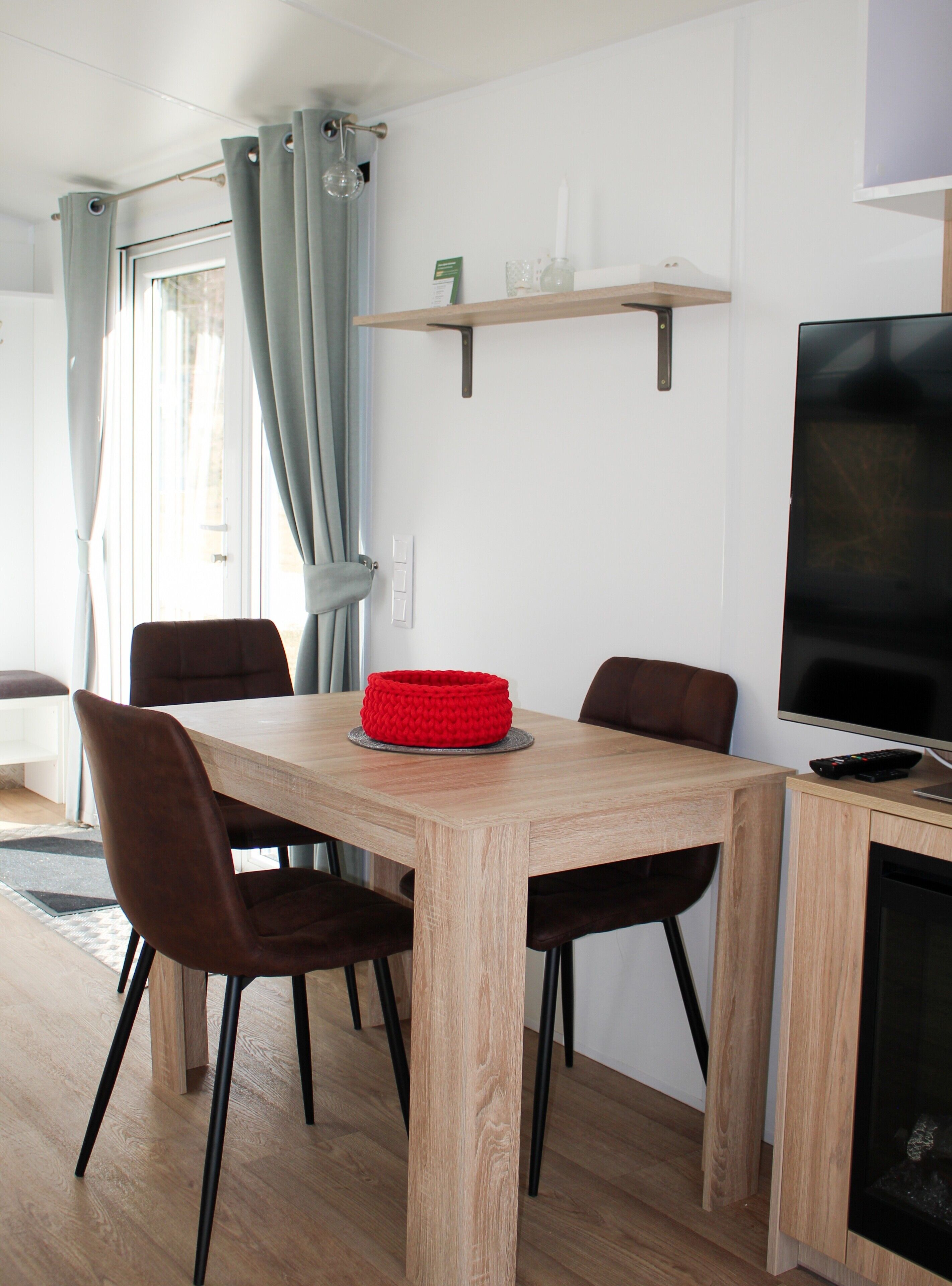 A dining area with a wooden table, four chairs, a red basket, a television, and a small shelf above the table.